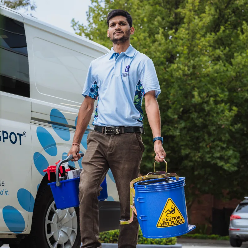 Man in uniform carrying cleaning supplies and a blue bucket with a 'Caution Wet Floor' sign near a service van.