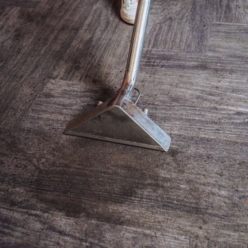 Metal carpet cleaning tool vacuuming a dark textured carpet with a person's white sneaker partially visible.