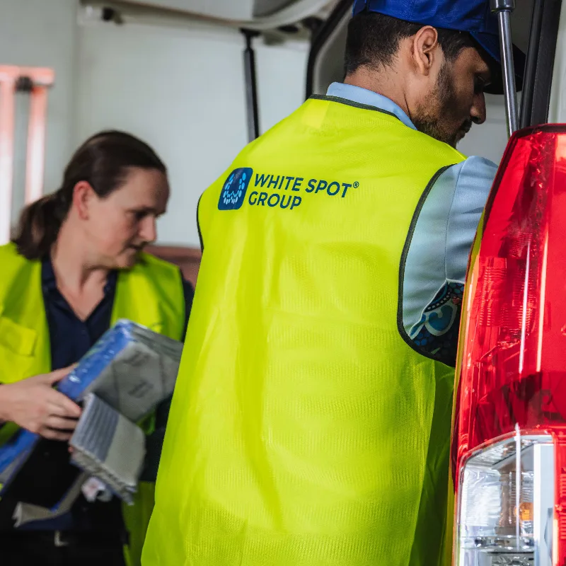 Two workers in yellow safety vests loading or unloading items from a vehicle, one vest has a White Spot Group logo.