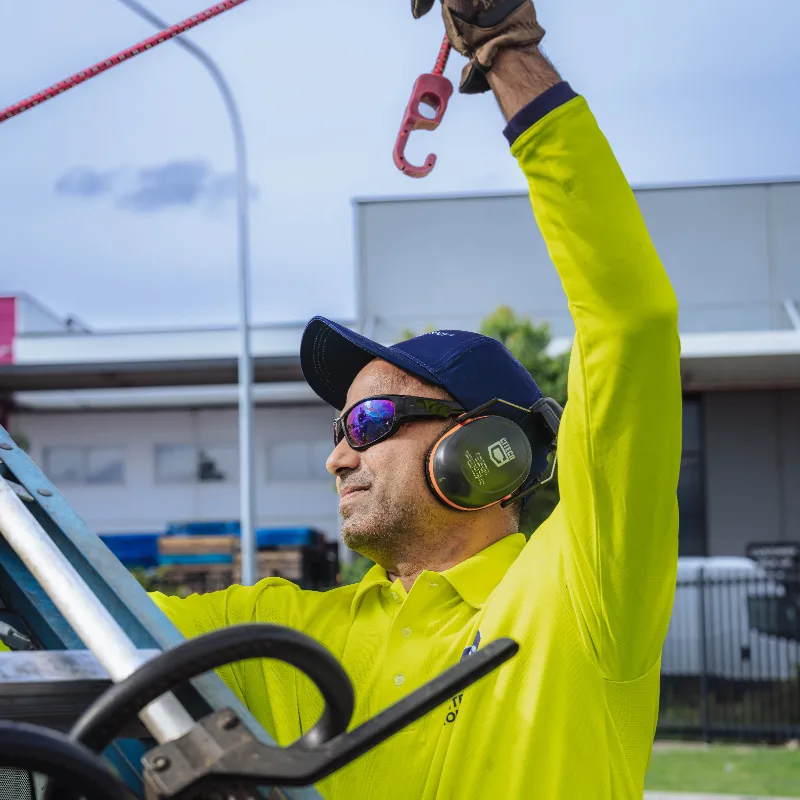 Worker in a yellow shirt and blue cap wearing safety glasses and earmuffs, securing a red hook while working outdoors.