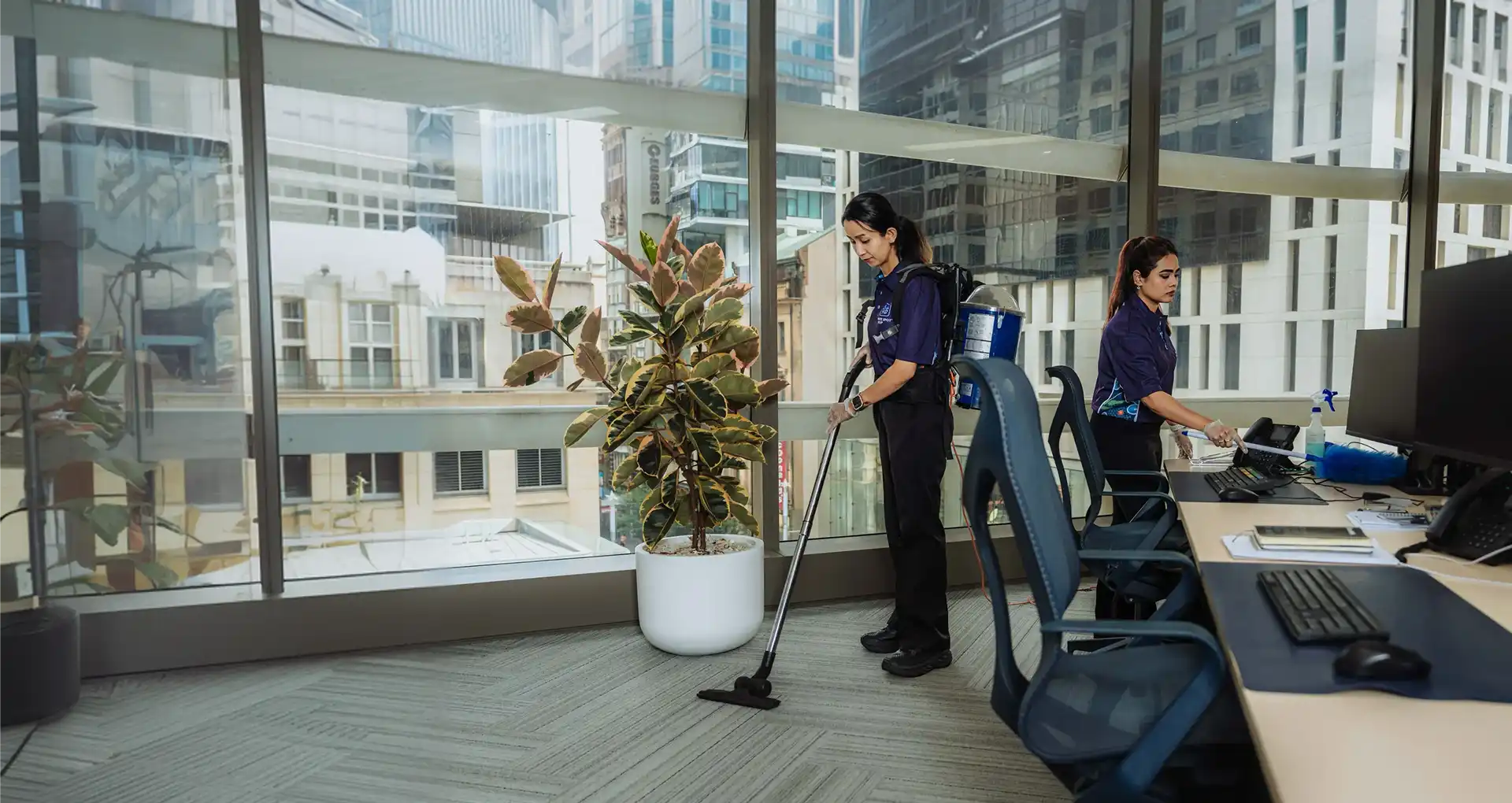 Two women cleaning an office space, one vacuuming the floor and the other wiping a desk near a computer.
