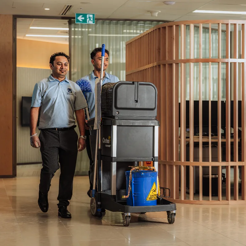 Two janitors walking down a hallway pushing a cleaning cart with supplies, including a caution wet floor bucket.