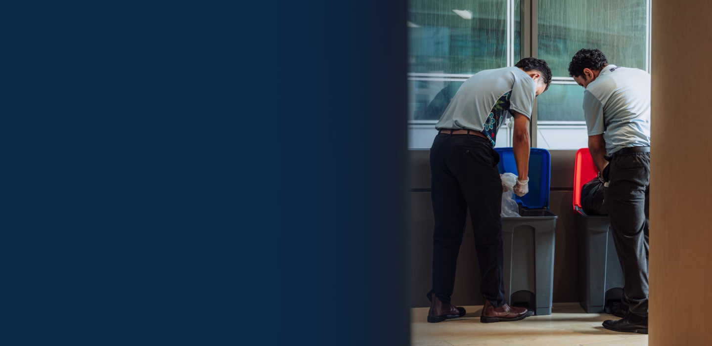 Two men in uniforms sorting waste into blue and red recycling bins indoors.