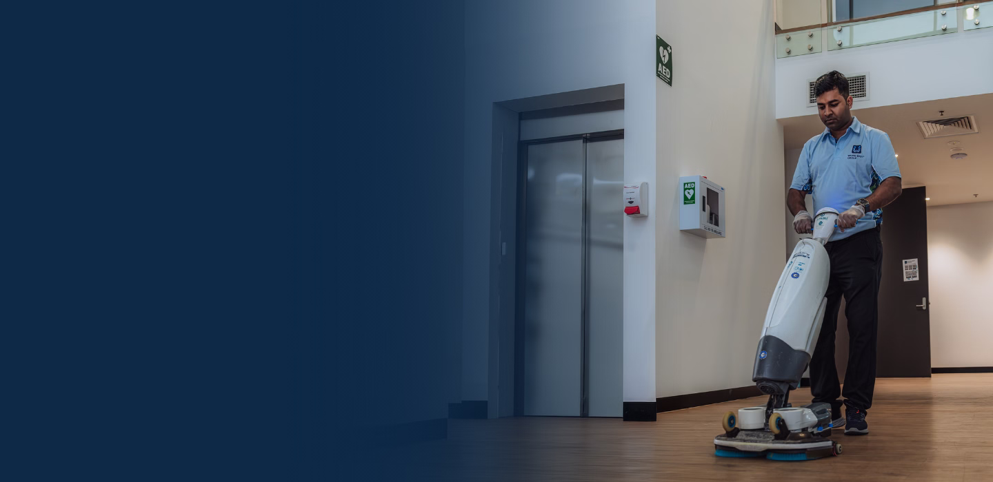 A man wearing gloves operates a floor cleaning machine on a wooden floor inside a modern building corridor.