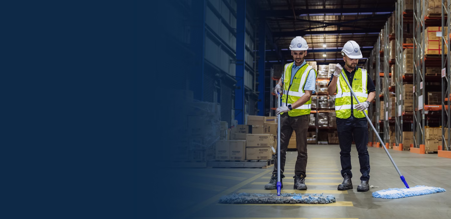 Two workers wearing hard hats and reflective vests cleaning a warehouse floor with large mops.