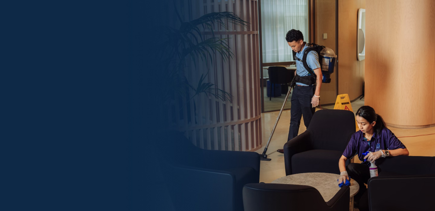 Two housekeeping staff cleaning a lounge area, one vacuuming the floor and the other wiping a table.