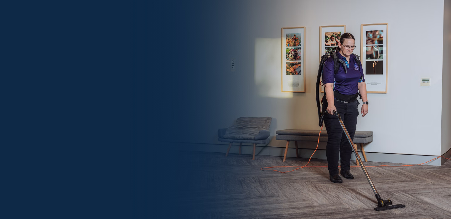 Woman wearing a backpack vacuum cleaner cleaning a room with gray flooring and framed pictures on the wall.