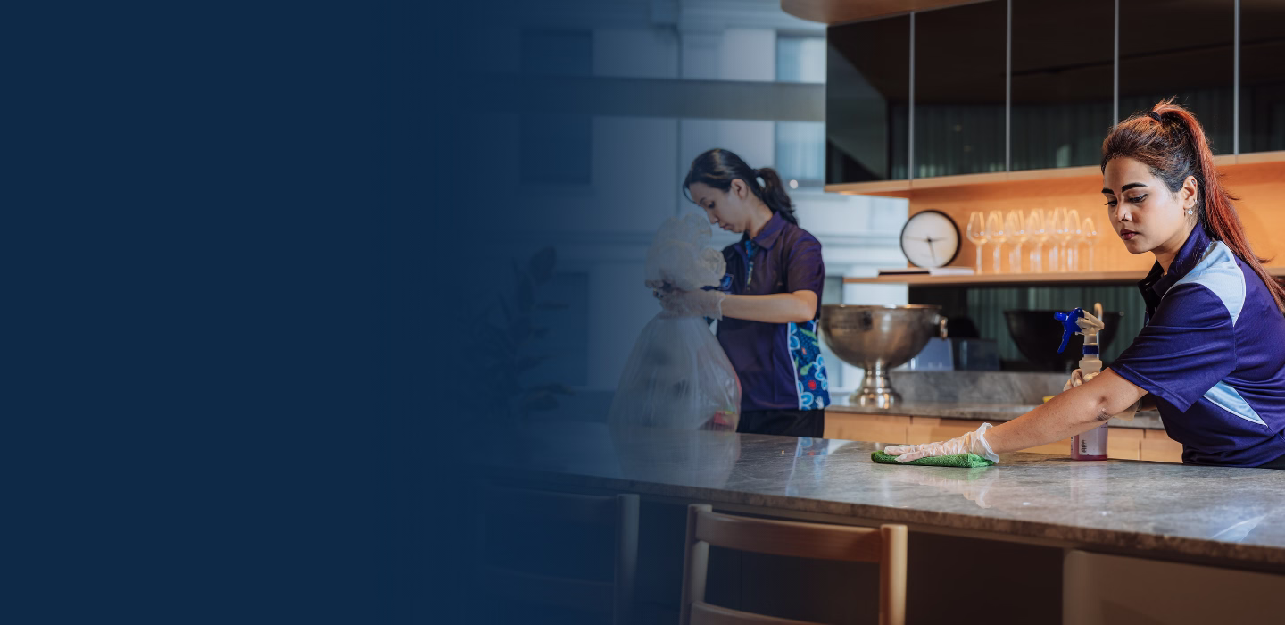 Two women wearing gloves cleaning a kitchen counter and handling a trash bag in a modern kitchen setting.