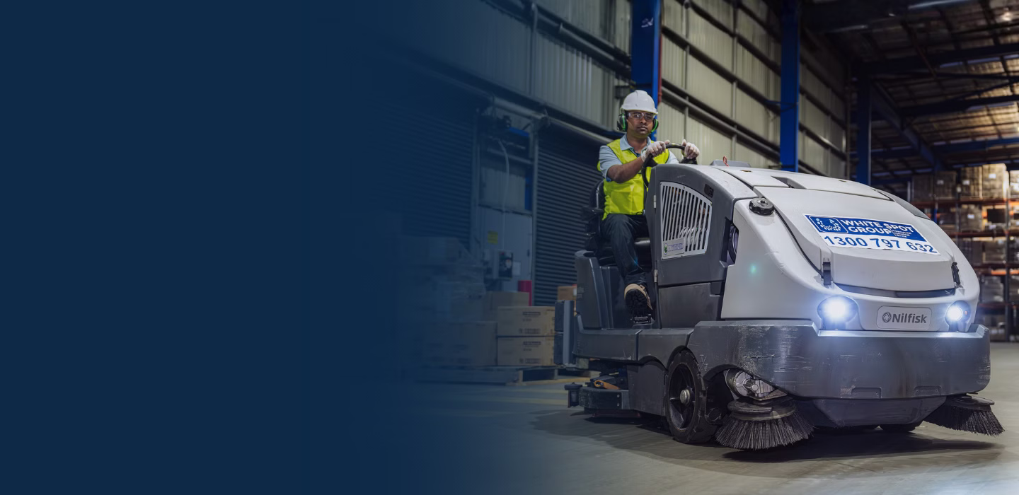 Worker wearing safety gear operating a Nilfisk floor cleaning machine inside a warehouse.
