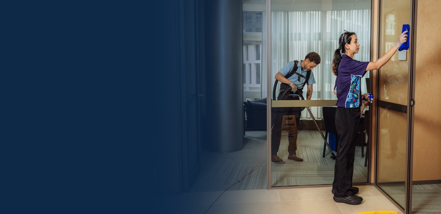 Two professional cleaners in uniforms cleaning an office space; one wiping a glass door and the other vacuuming carpet.