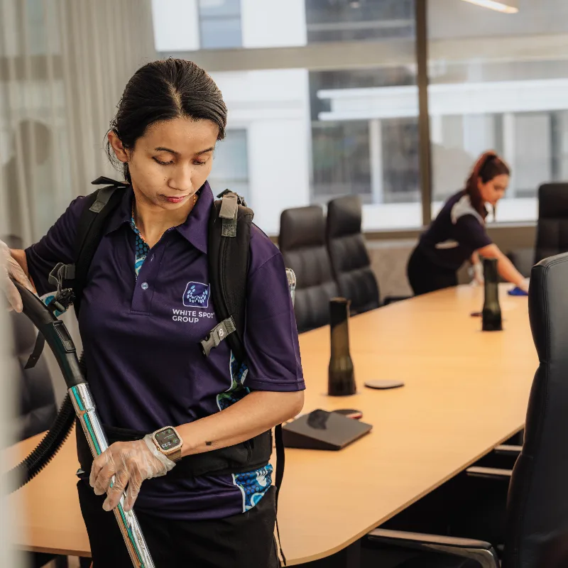 Two professional cleaners wearing purple White Spot Group shirts cleaning a modern office conference room with black leather chairs.
