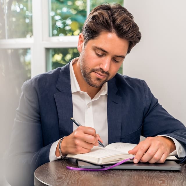 Man in a navy blazer sitting at a table writing in a notebook, with a window in the background.