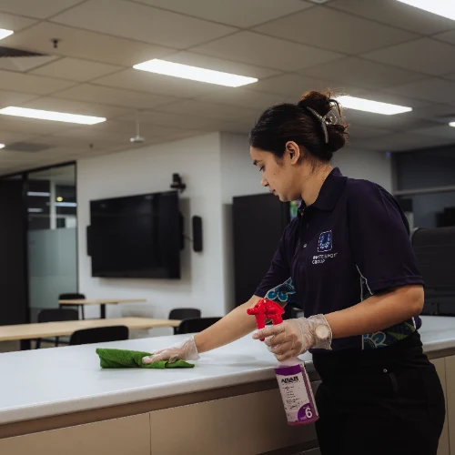 Woman wearing gloves cleaning a white countertop with a green cloth and spray bottle in a modern office setting.
