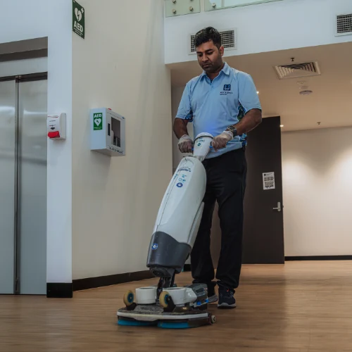 Man wearing blue shirt and gloves operating a floor cleaning machine in a hallway with wooden flooring.
