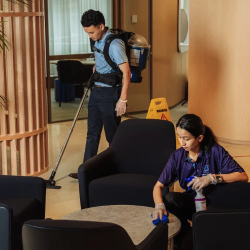 A man vacuuming a hotel lobby floor and a woman cleaning a table, both wearing gloves and cleaning uniforms.