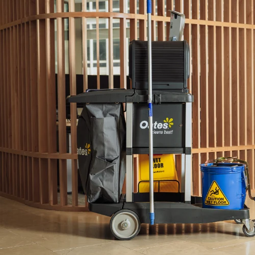 A black cleaning cart with a mop, a trash bag, a caution wet floor sign, and a blue bucket inside a modern wooden grid room.