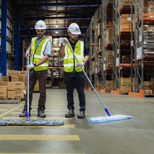 Two warehouse workers wearing safety helmets and yellow reflective vests cleaning the floor with large mops.