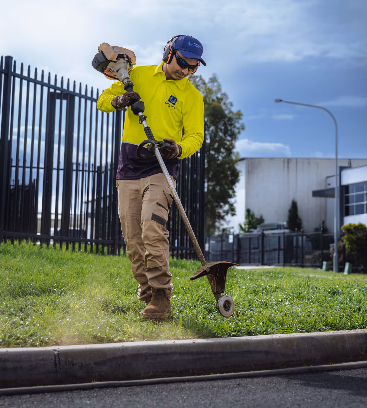 Worker wearing protective gear using a grass trimmer to edge a lawn near a sidewalk in an urban area.