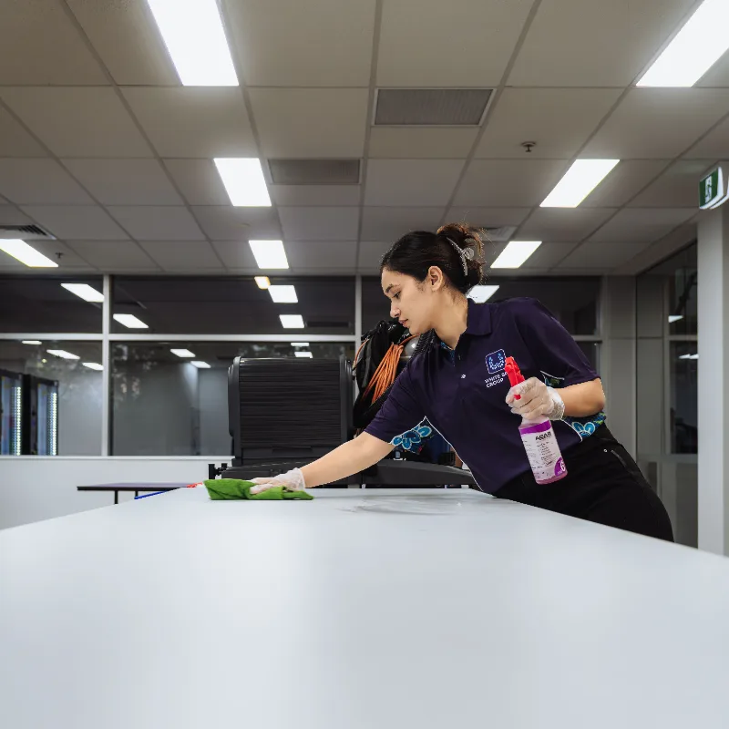 Woman in uniform cleaning a white table with a green cloth and spray bottle in a well-lit modern office.
