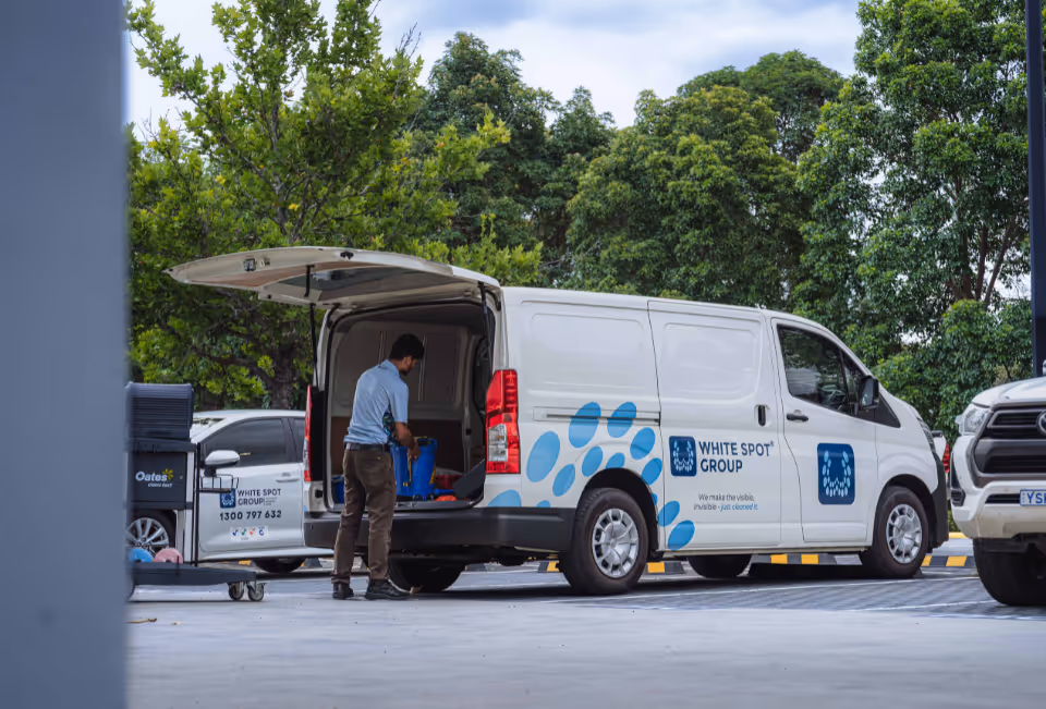 Man loading cleaning supplies into the back of a White Spot Group branded white van parked outdoors near trees.