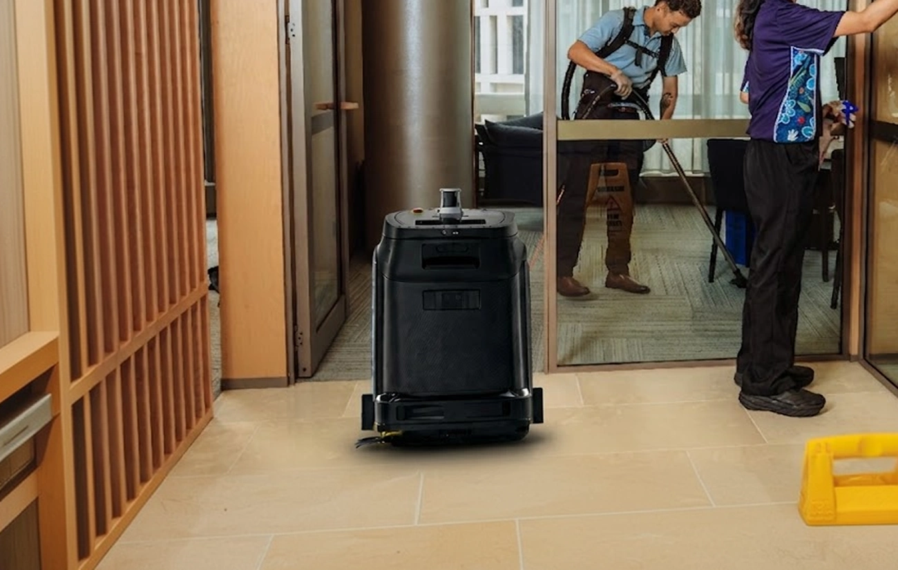 Robot vacuum cleaner in hallway with two people cleaning inside office with glass doors.