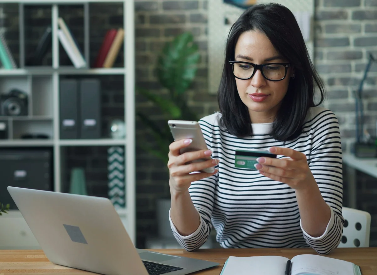 Une femme lit un message sur son téléphone, à son bureau devant son ordinateur portable