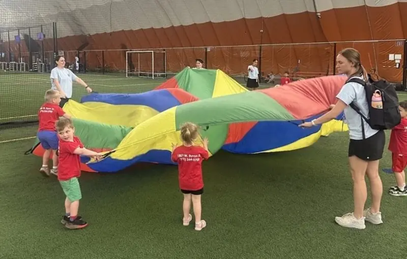 Children in Black Bear Academy shirts play a parachute game with teachers on an indoor turf field
