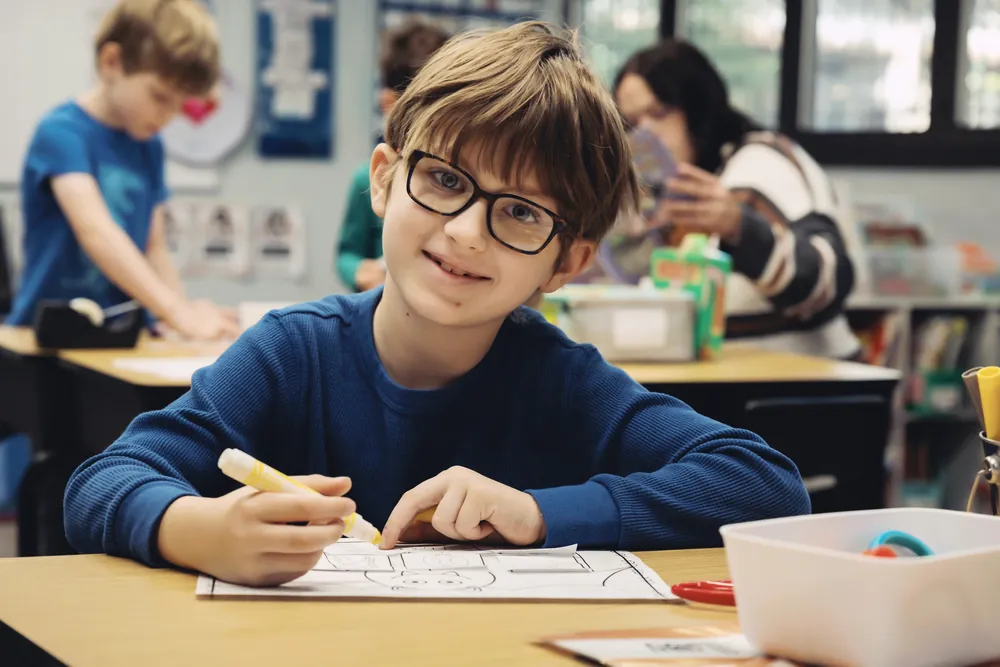 A Black Bear Academy student smiles while working on a drawing activity at his desk