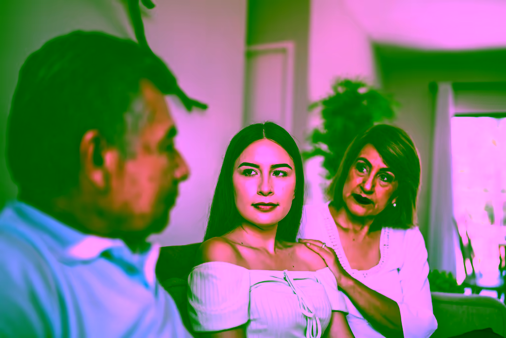 A young woman and an older woman sit side by side on a sofa, the older woman resting a hand on the young woman's shoulder, while a man in the foreground looks away.