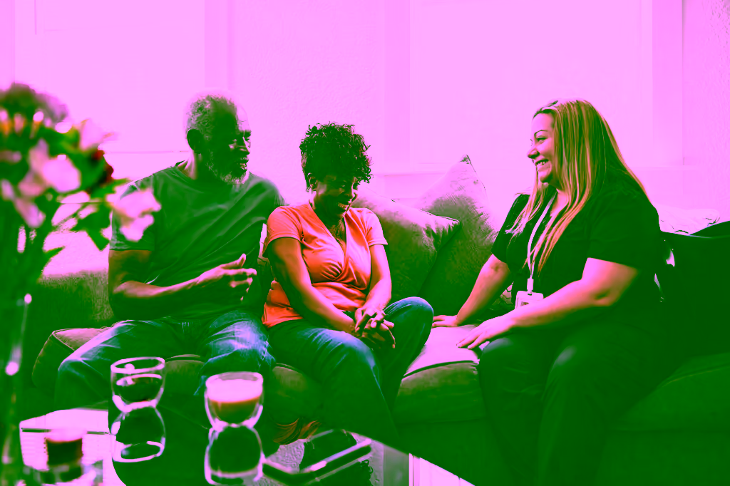 A smiling woman with an ID badge sits on a sofa and talks with an older man and woman seated beside her.