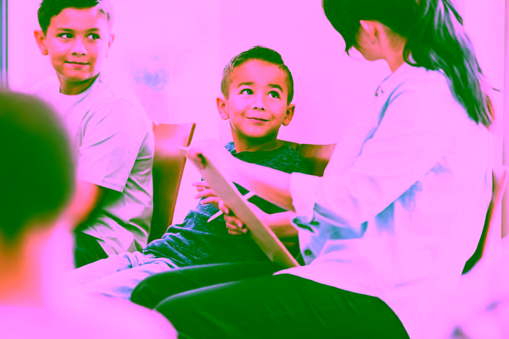A young boy looks up and smiles at a woman holding a clipboard while another boy sits beside him in a group setting.