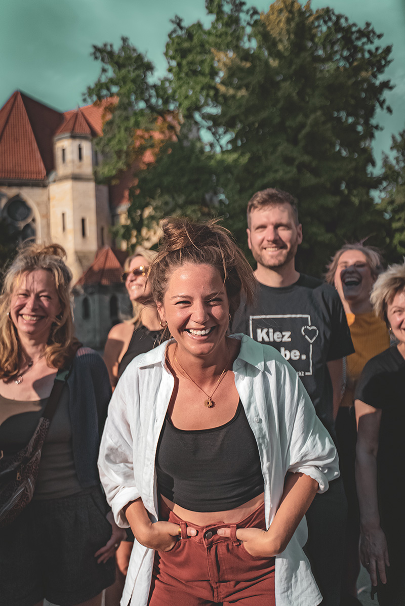 Alina Zimmermann steht lächelnd mit den Händen in den Hosentaschen vor einer Gruppe von Menschen nahe einem historischen Steingebäude.
