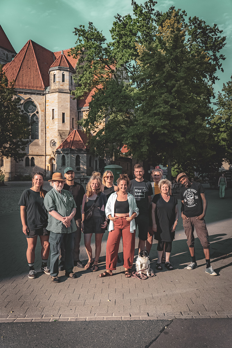 Alina Zimmermann steht mit acht weiteren Menschen und einem Hund vor einem historischen Gebäude und Bäumen an einem sonnigen Tag.