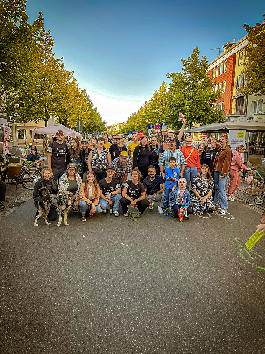 Eine größere Gruppe von Menschen posiert mit zwei Hunden auf einer von Bäumen und Wohnhäusern gesäumten Straße.