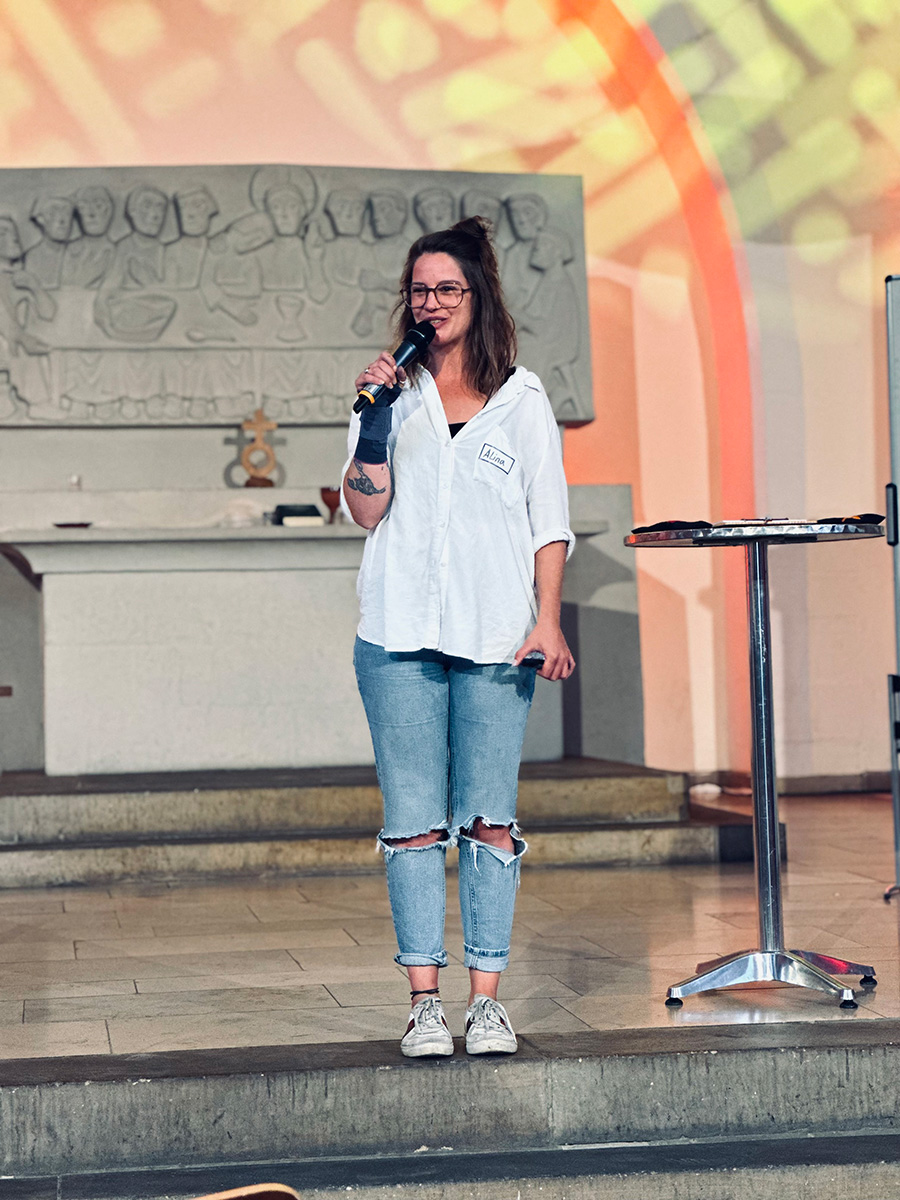 Alina Zimmermann spricht mit einem Mikrofon auf einer Bühne; im Hintergrund ist ein Relief im Innenraum einer Kirche zu sehen.