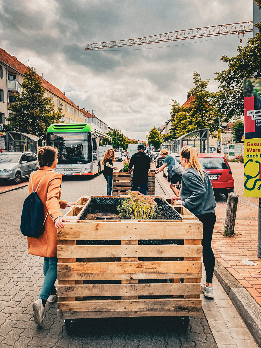 Alina Zimmermann und vier weitere Personen schieben bepflanzte Holzkästen über eine Stadtstraße mit Autos und einem grünen Bus im Hintergrund.