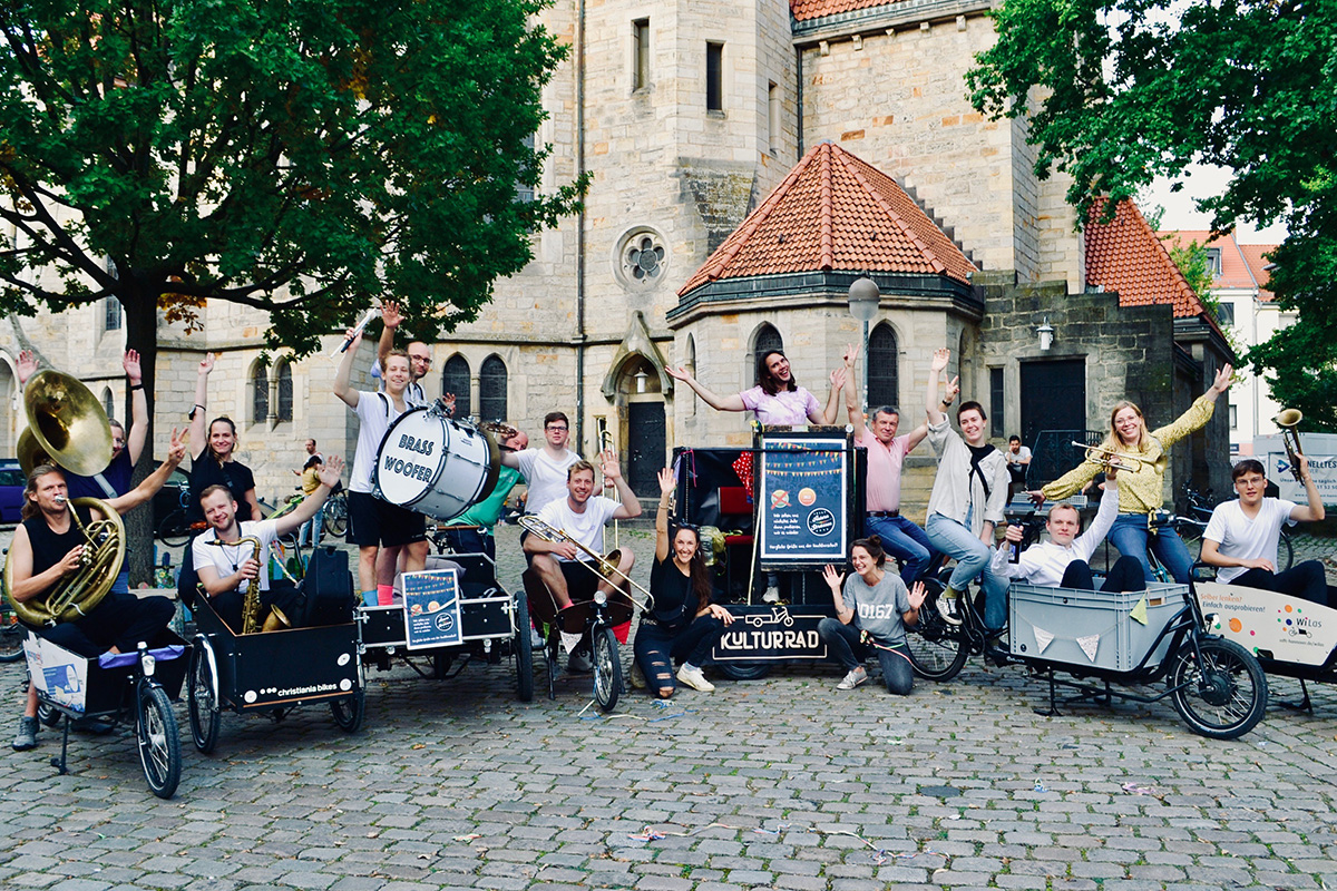Alina Zimmermann und eine Gruppe von Musiker:innen mit Blasinstrumenten posieren mit geschmückten Lastenrädern vor einer Kirche.