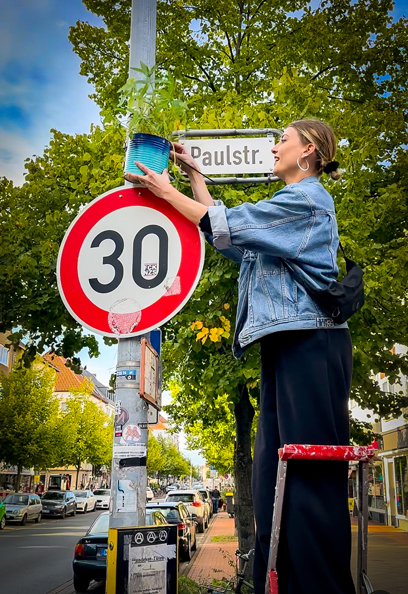 Eine Person auf einer Leiter befestigt einen blauen Pflanztopf mit Grünpflanzen an einem Laternenmast neben einem Tempo-30-Schild.