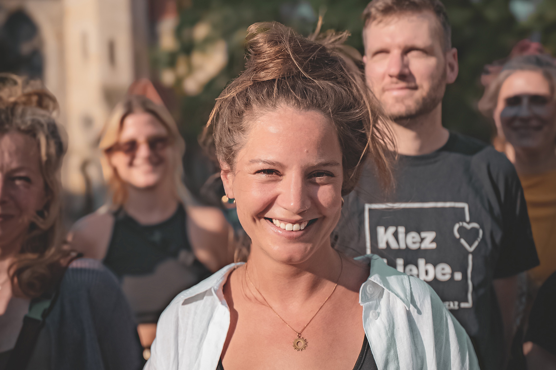 Alina Zimmermann lächelt im Freien in die Kamera, hinter ihr steht eine Gruppe von Menschen in der Sonne.