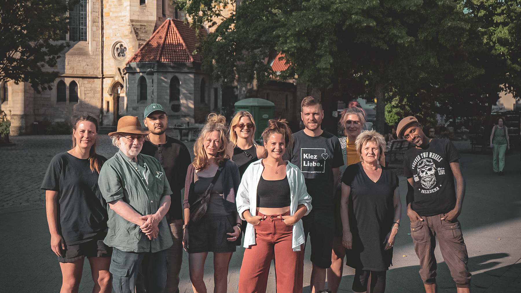 Alina Zimmermann lächelt im Freien in die Kamera, hinter ihr steht eine Gruppe von Menschen in der Sonne.