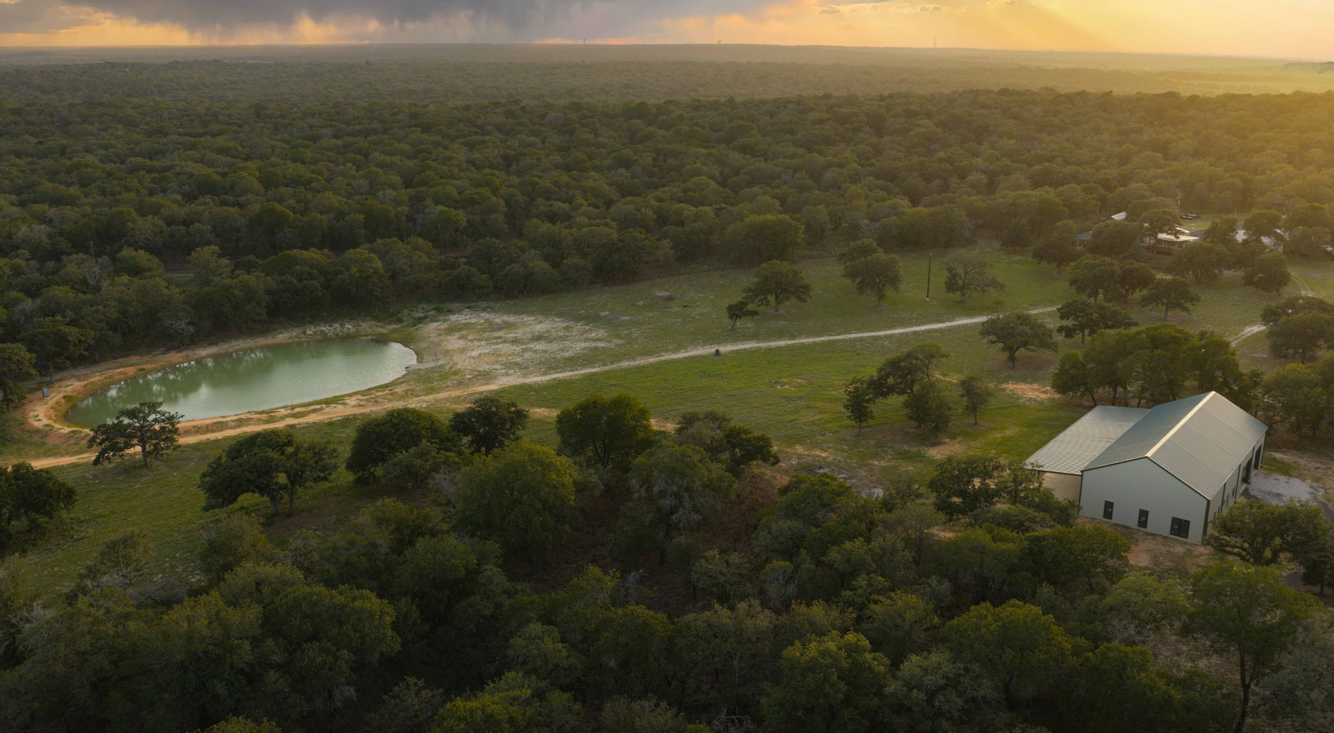 Aerial view of rural property with white barn, pond, and dense forest at golden hour