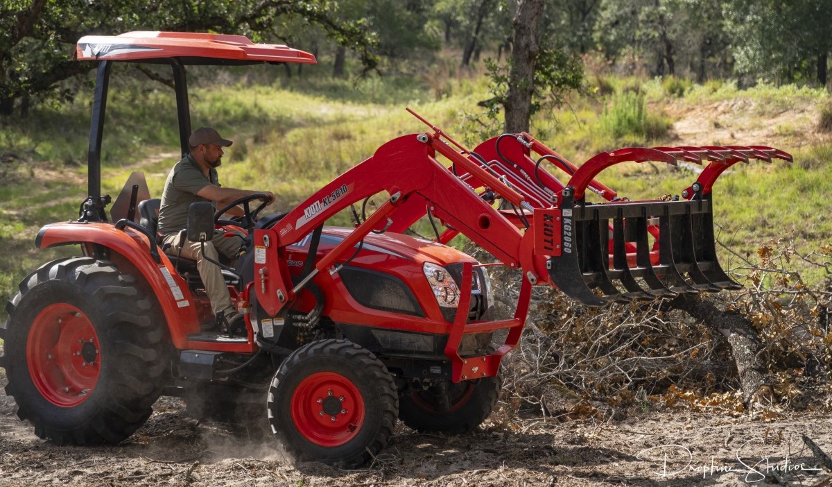 Farmer operating red compact tractor with front-end loader and attachment in wooded terrain.