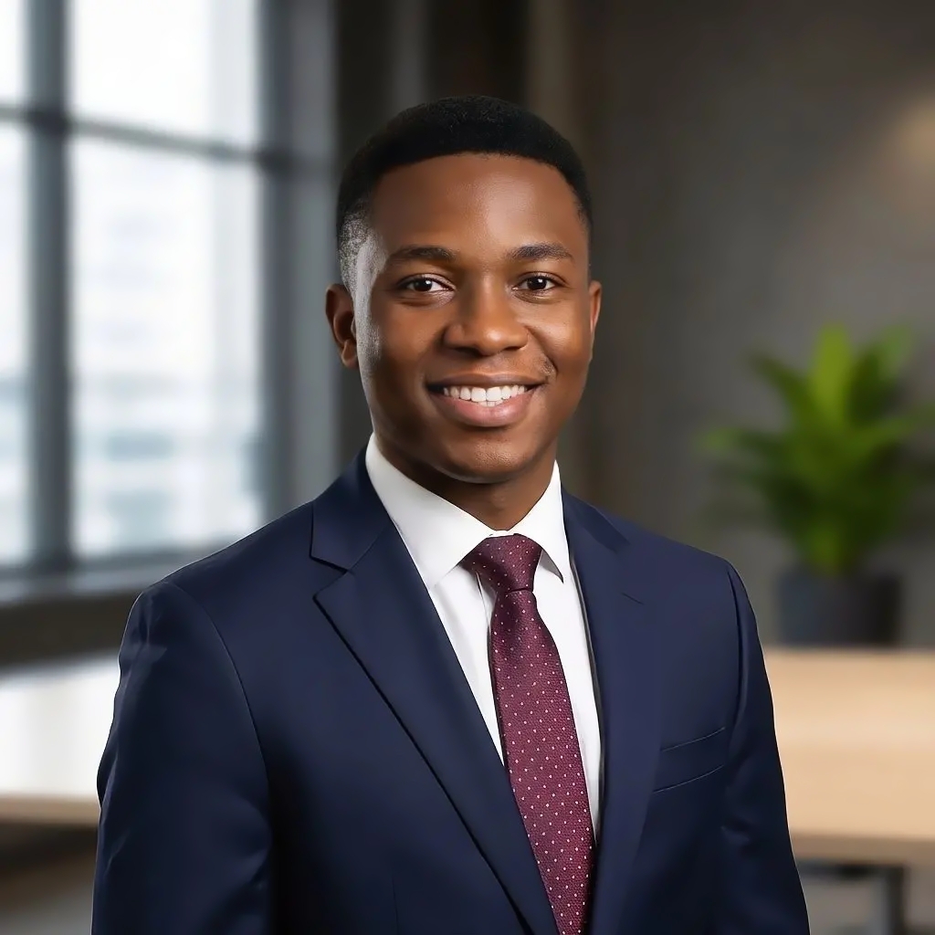 Smiling young man wearing a navy blue suit, white shirt, and maroon polka dot tie, standing in a modern office with a blurred window and plant in the background.