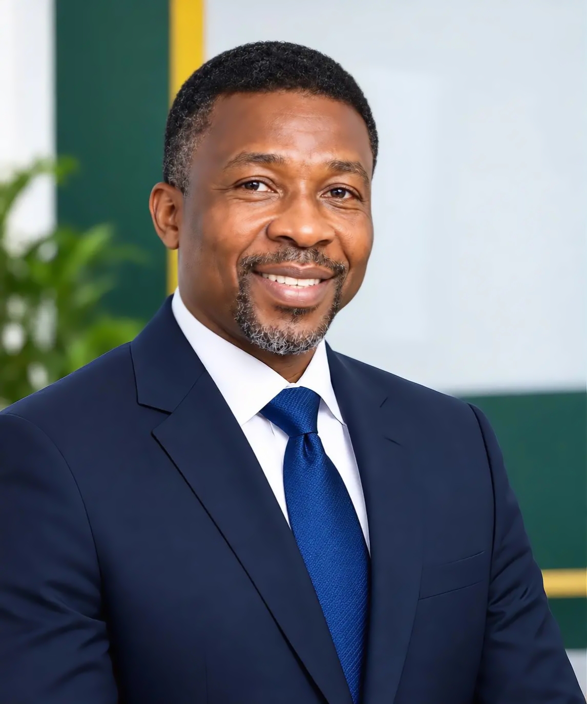 Smiling man with short curly hair and a trimmed salt-and-pepper beard wearing a navy blue suit, white shirt, and blue tie, posed indoors with a blurred background featuring green and yellow accents and a plant.