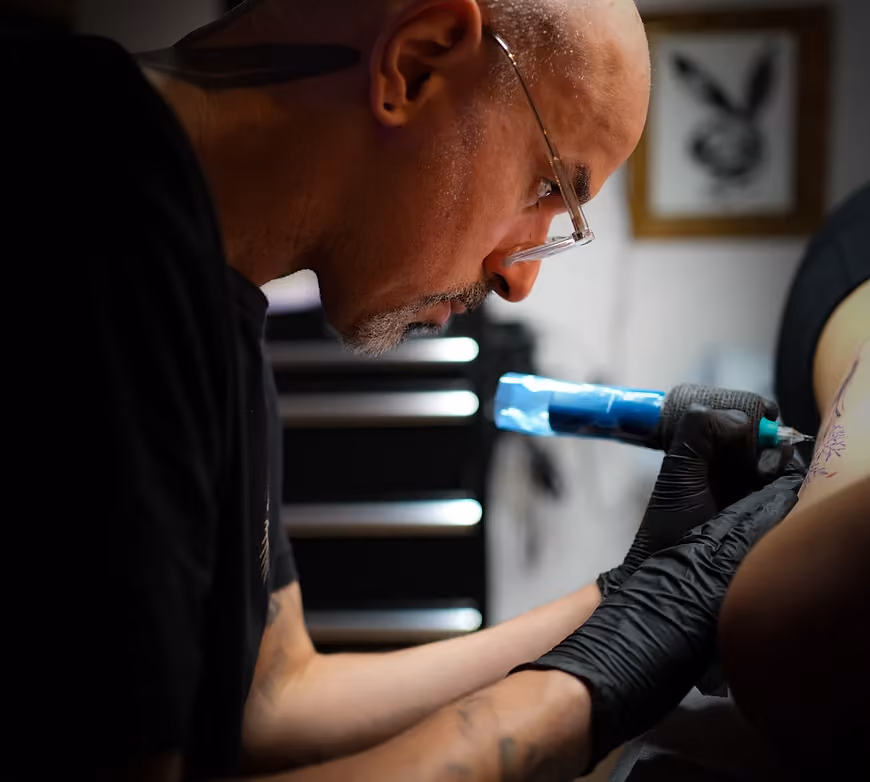Tattoo artist wearing glasses and black gloves focused on tattooing a person's upper arm in a studio setting.