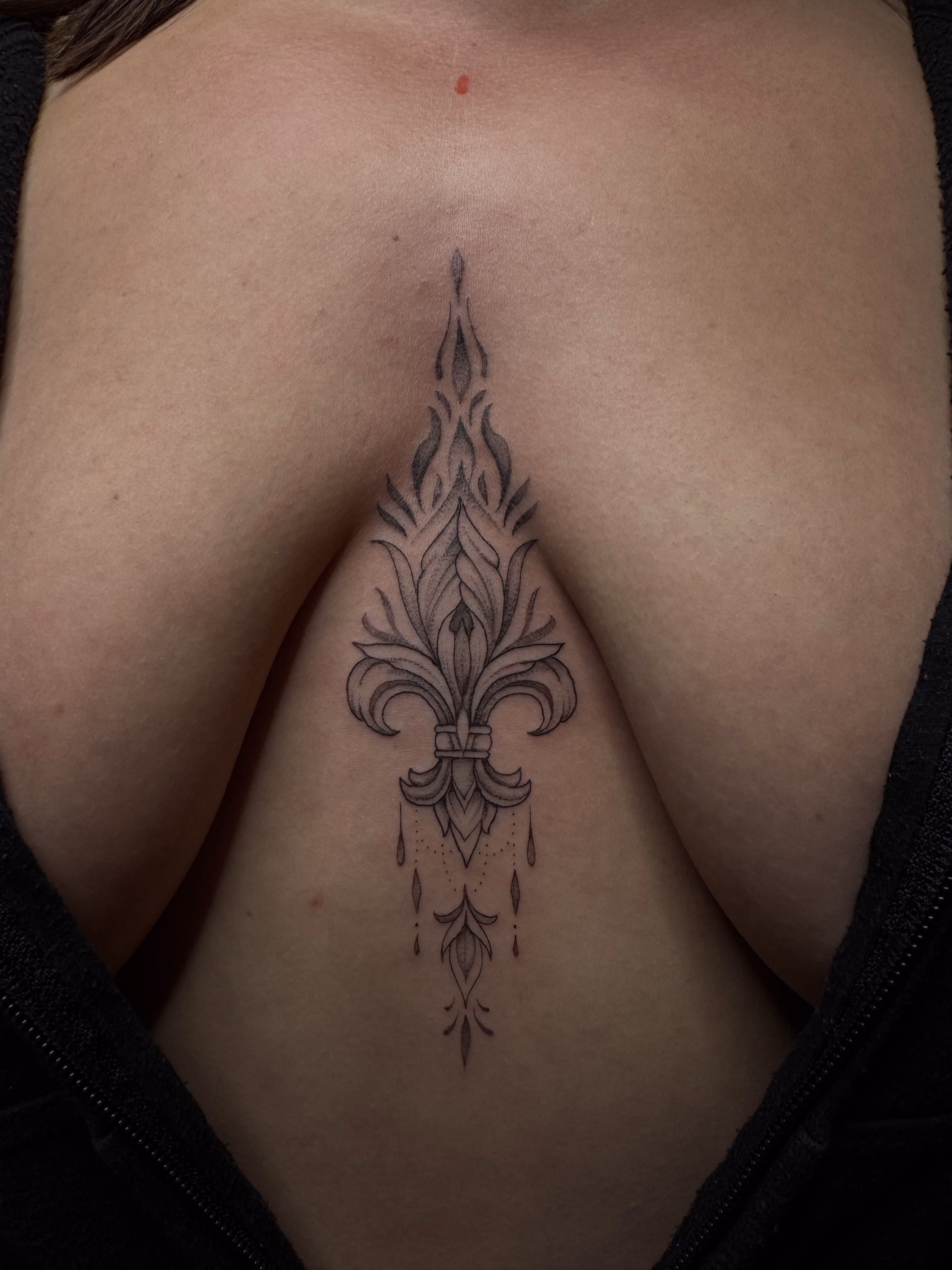 Intricate black ink tattoo of a stylized fleur-de-lis with ornamental details and small teardrop shapes on the cleavage area between the breasts.