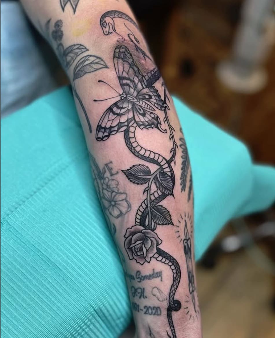 Forearm tattoo featuring a large black and gray butterfly perched on a twisting vine with leaves and a rose, surrounded by smaller floral and candle tattoos and cursive text.