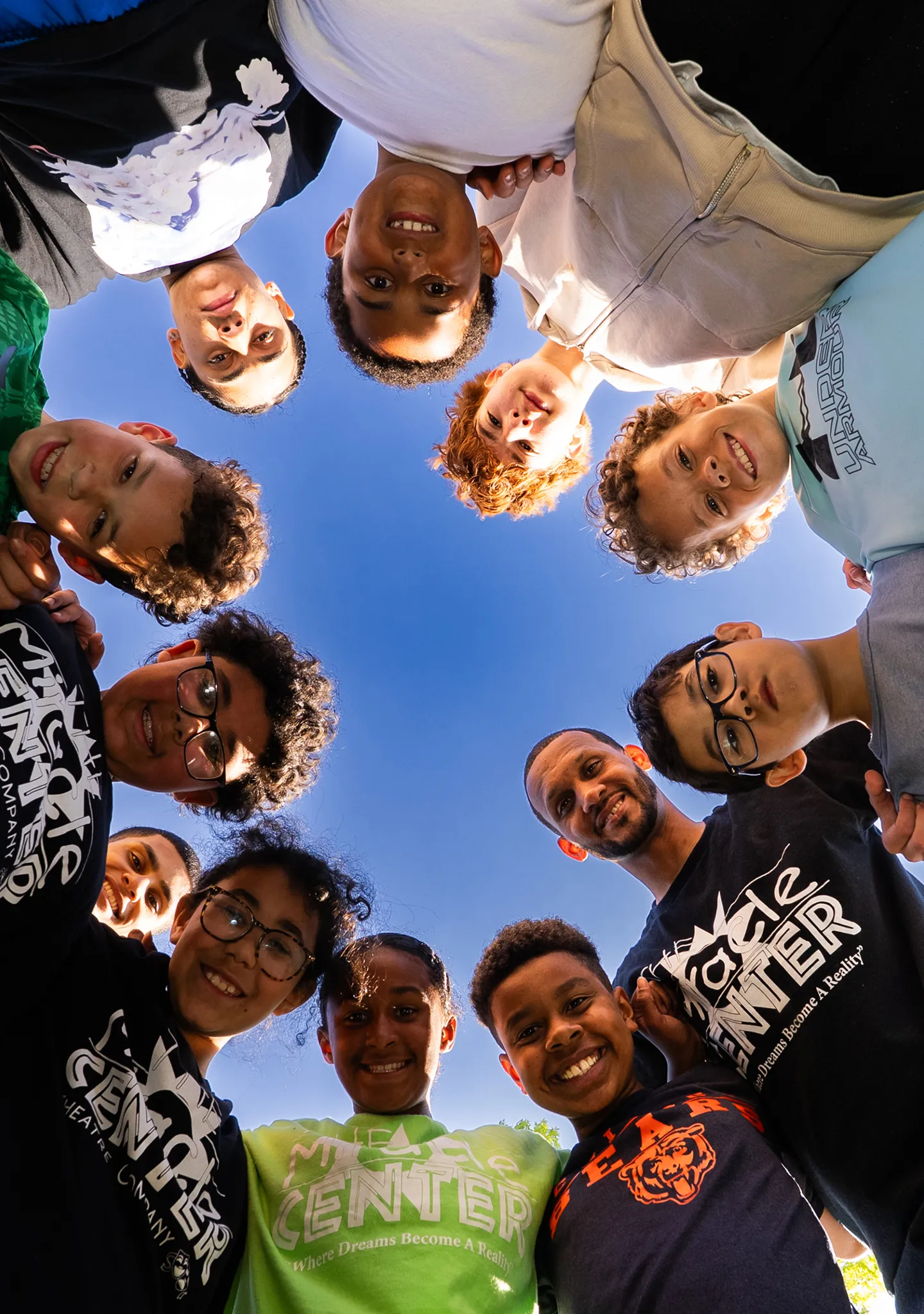 A diverse group of smiling children and one adult form a circle, looking down at the camera against a clear blue sky.