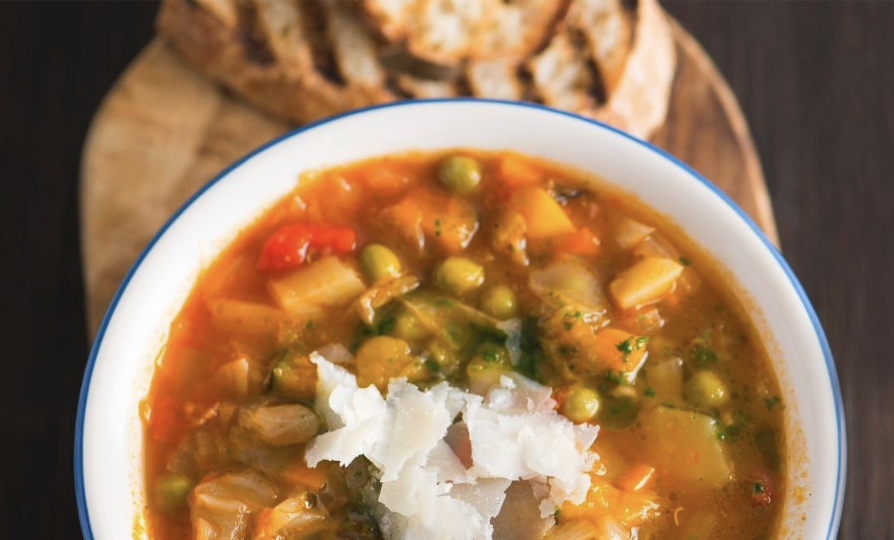 A bowl of vegetable soup topped with shaved cheese, served with grilled bread