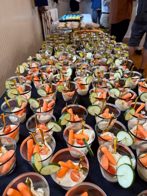 Rows of cups filled with hummus topped with baby carrots, cucumber slices, and cherry tomato halves on a table.